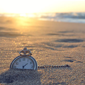 pocket watch stuck in the sand of a beach with sun setting on the ocean in the background